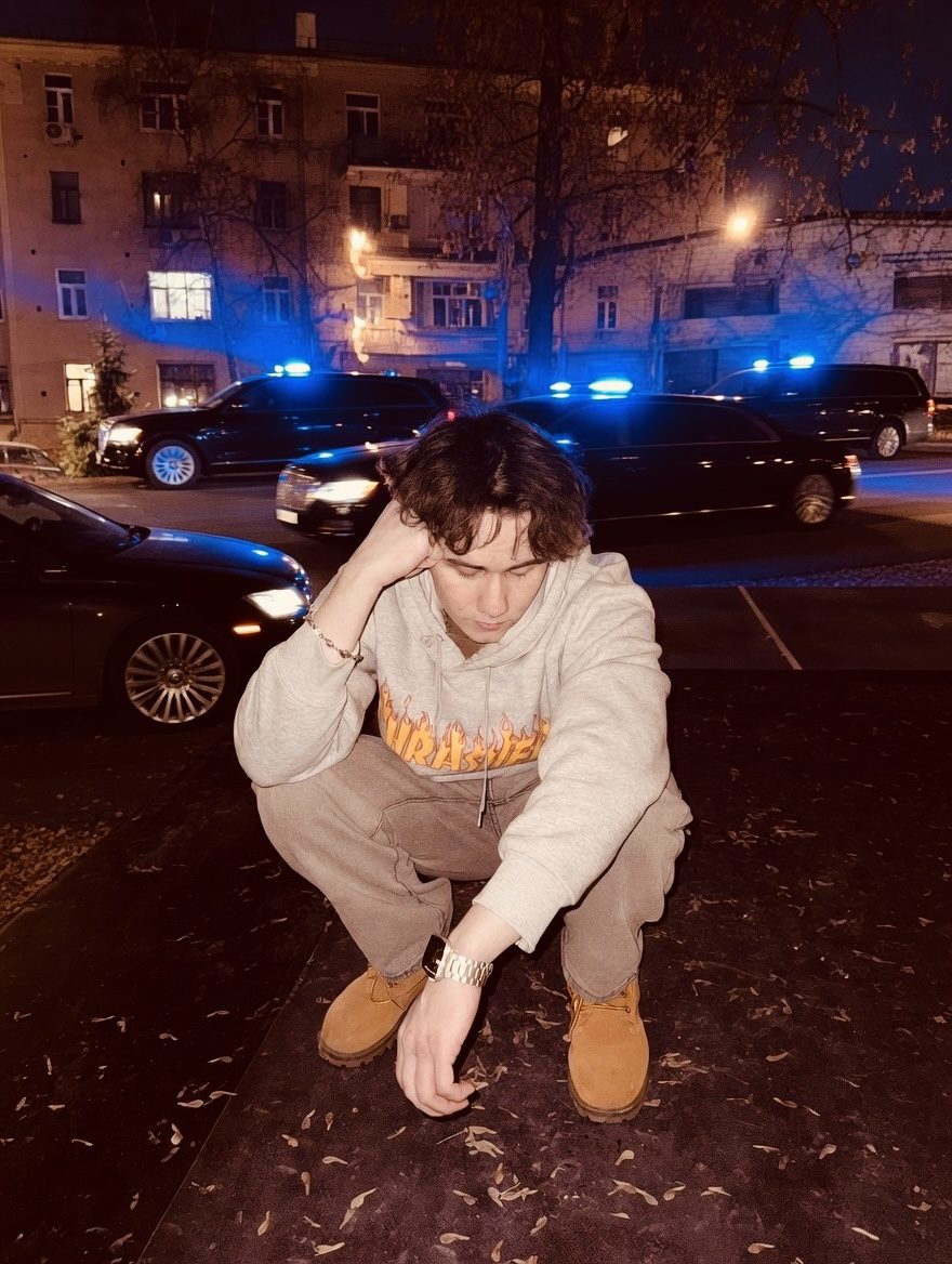 Ярослав Танго сидит на бордюре ночью в худи Thrasher, за спиной синие мигалки